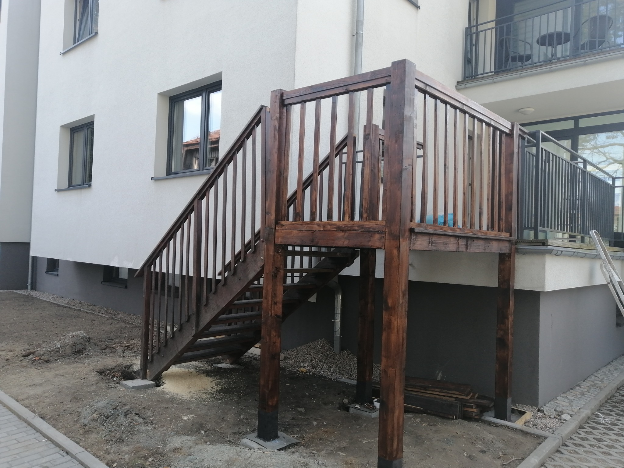 Drewniana konstrukcja schodów zewnętrznych z balustradą, prowadząca do drzwi na piętrze budynku z białymi ścianami i szarym cokołem, widoczne ślady prac ziemnych wokół.