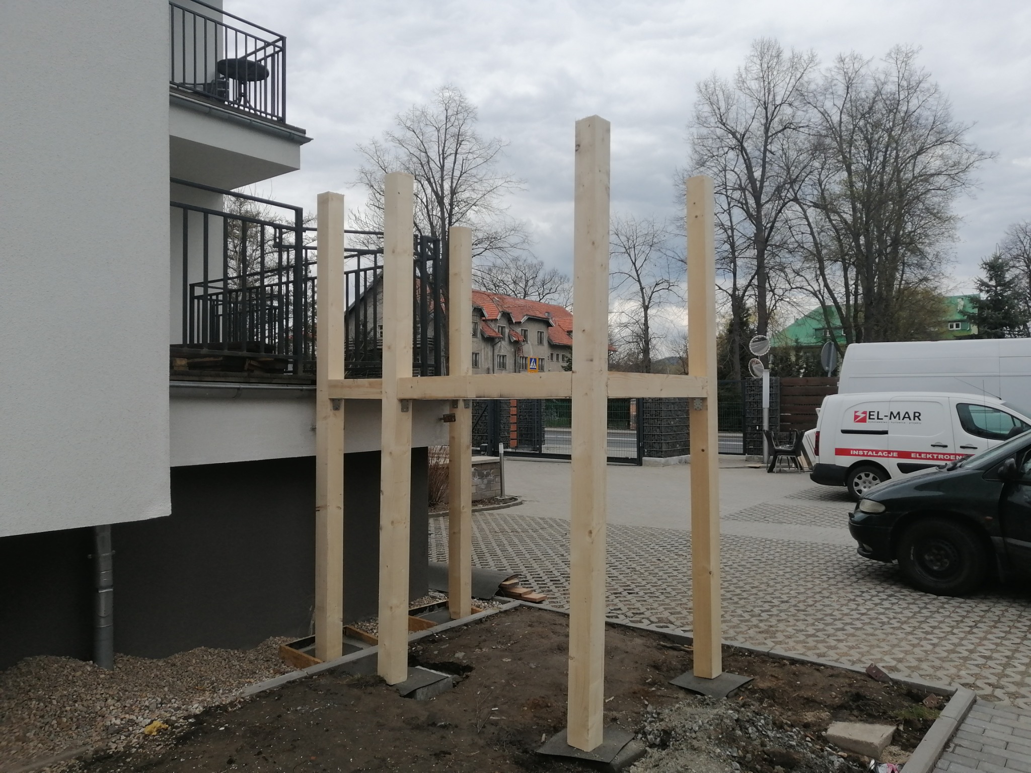 Surowa drewniana konstrukcja pergoli w trakcie montażu, widoczne słupy i belki poprzeczne, na tle budynku mieszkalnego i zaparkowanego samochodu dostawczego.