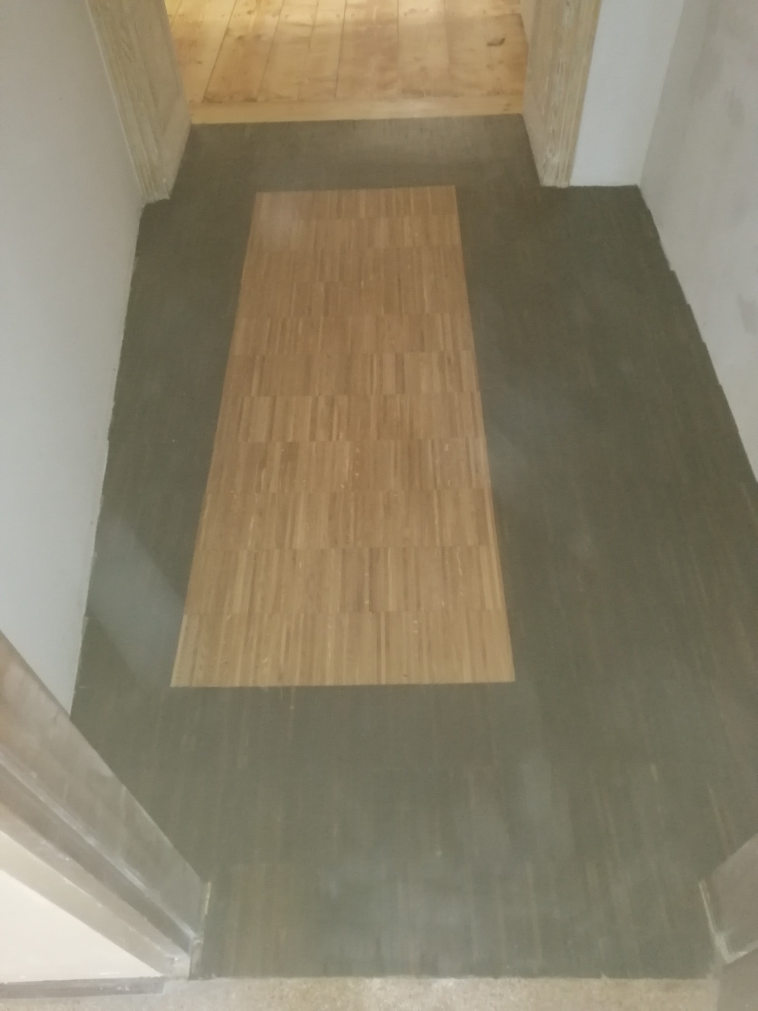 Fragment podłogi w korytarzu z ułożonym parkietem w centralnej części, otoczonym szarą wylewką wyrównującą, widok z góry na etapie prac wykończeniowych.