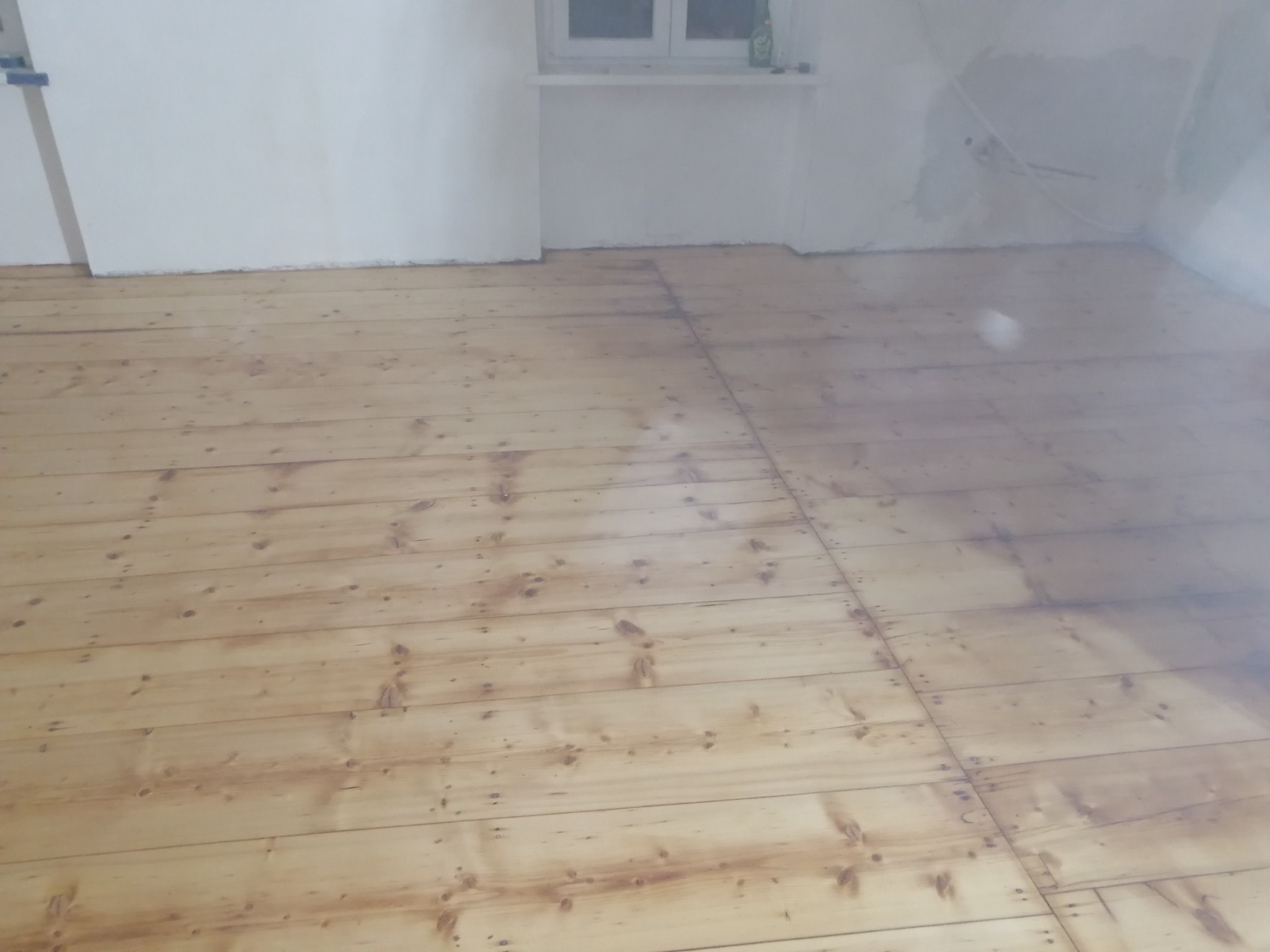 Wypolerowana podłoga z desek w jasnym odcieniu, widoczne ślady po szpachlowaniu i gwoździach, świeżo po nałożeniu lakieru, ściany w tle przygotowane do malowania.