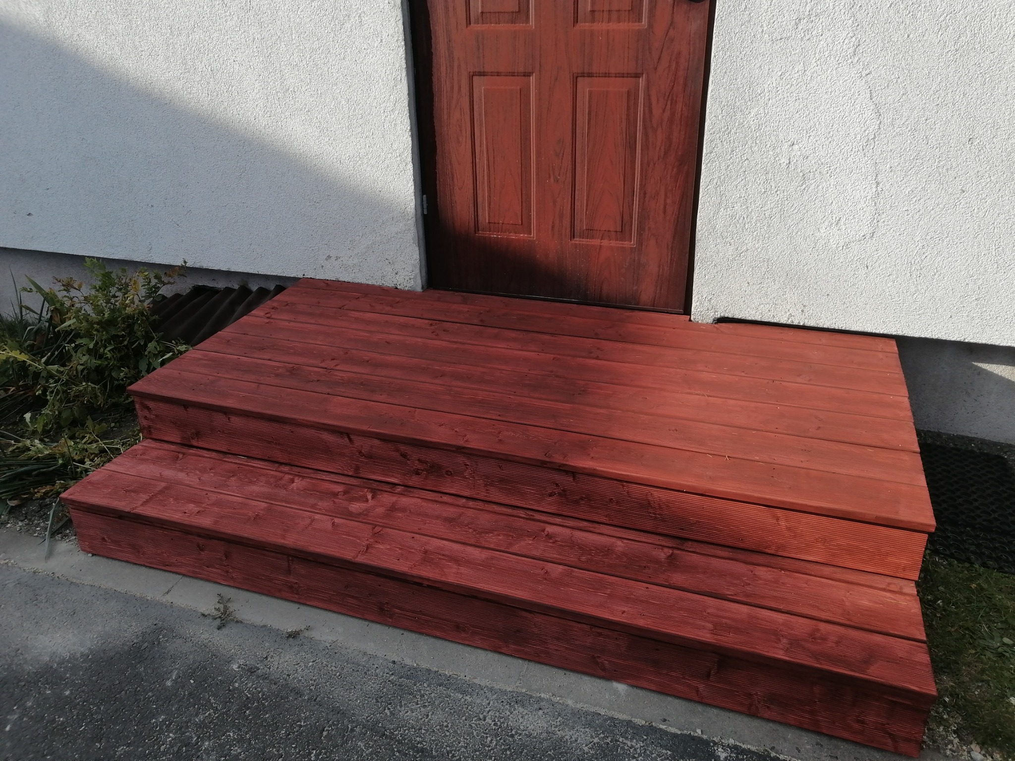 Czerwone, drewniane schody wejściowe prowadzące do drzwi w jasnym budynku, widoczne z poziomu gruntu.