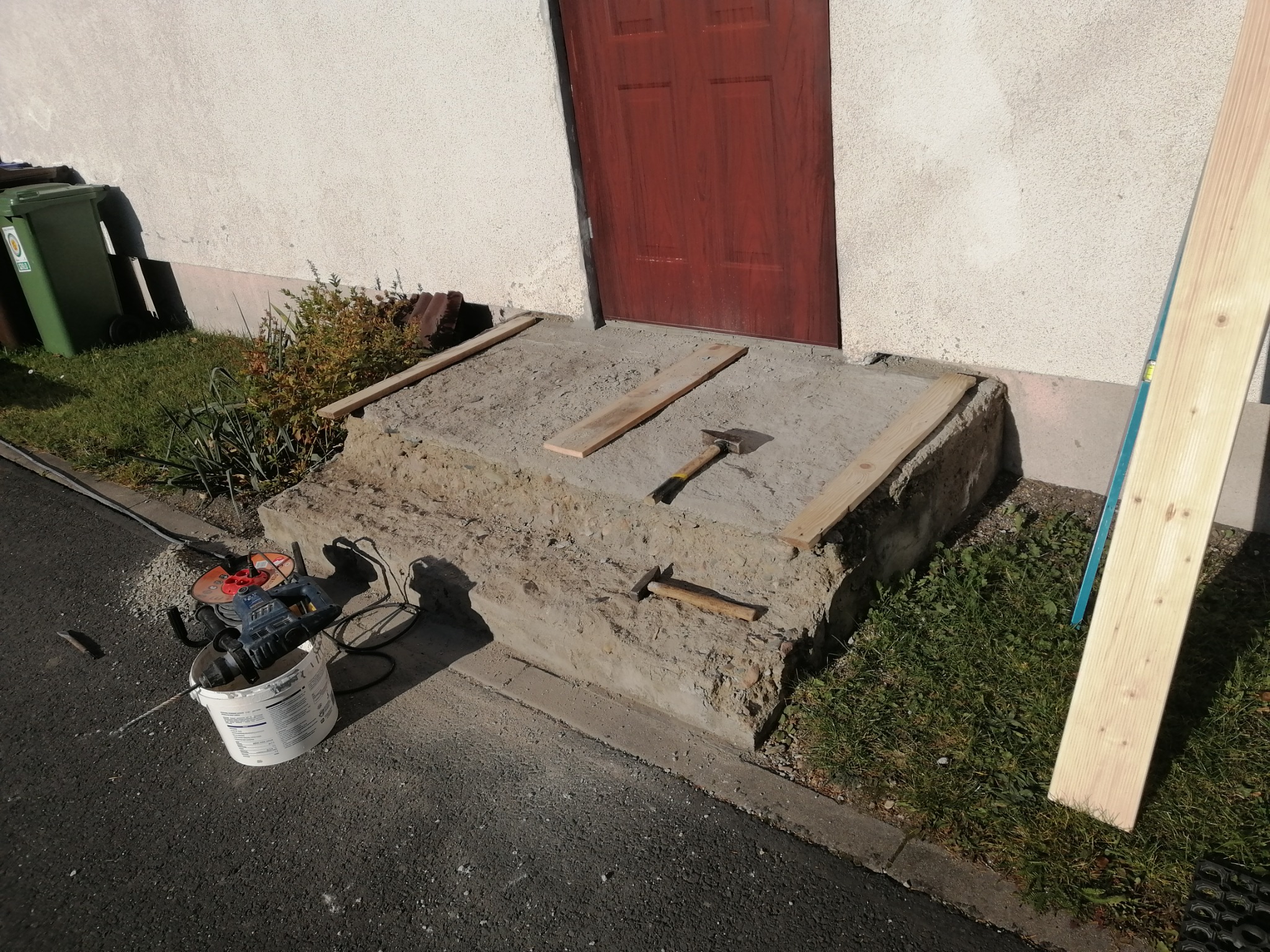 Betonowe schody wejściowe do domu w trakcie naprawy, widoczne narzędzia, deski szalunkowe i młotek.
