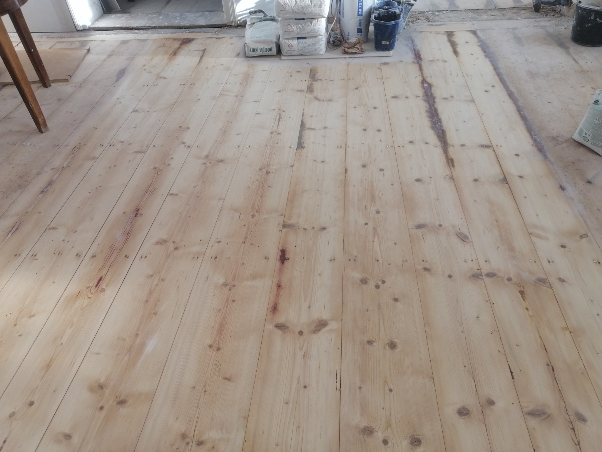 Surowa, jasna podłoga z desek sosnowych, częściowo przeszlifowana, z widocznymi śladami po starym wykończeniu i wystającymi gwoździami, przygotowana do dalszej obróbki, worki z zaprawą w tle.