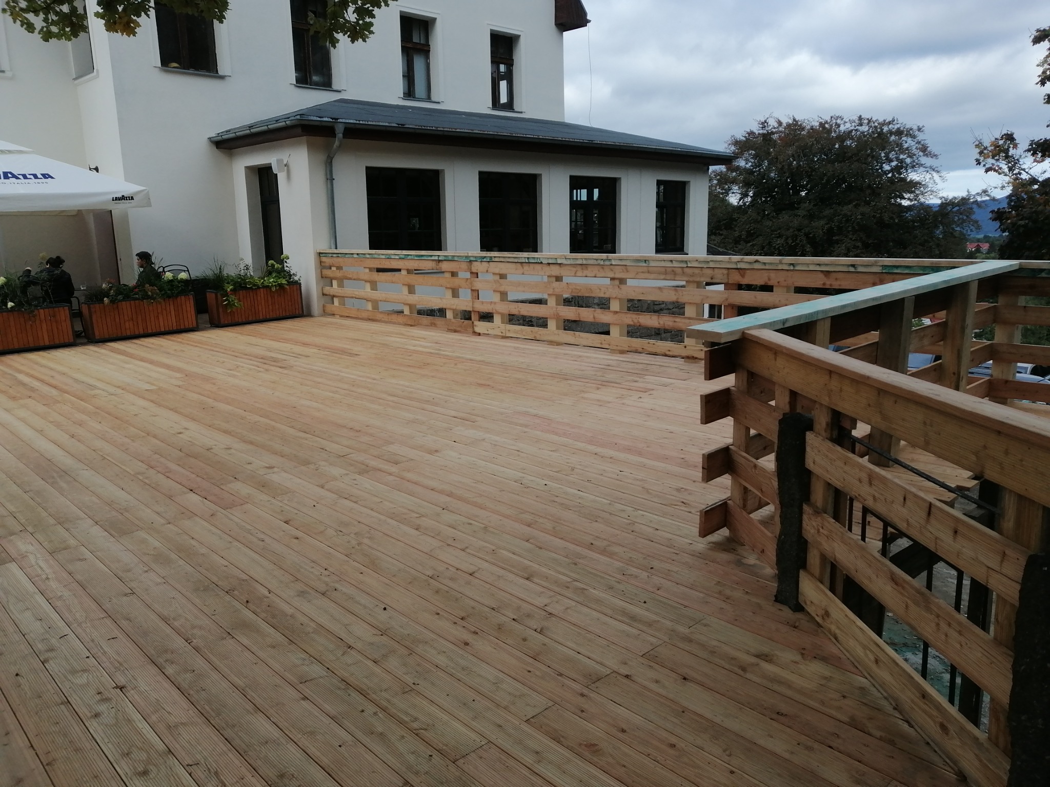 Nowo wybudowany drewniany taras z balustradą przy białym budynku, widoczne donice z roślinami i parasol kawiarniany w tle.