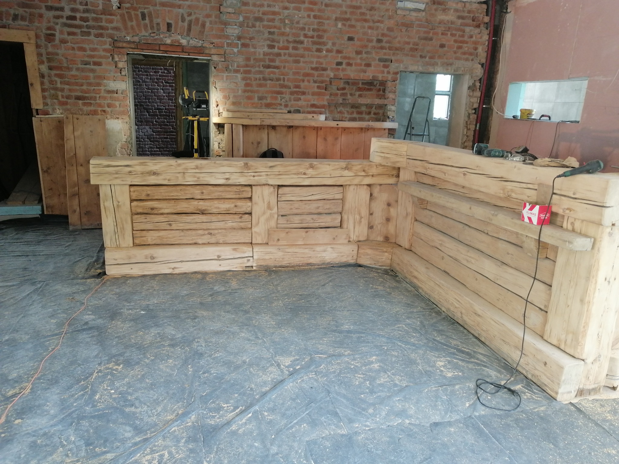 Surowa drewniana konstrukcja baru z bali w trakcie montażu, widoczne narzędzia, odkryta cegła, podłoga zabezpieczona folią.