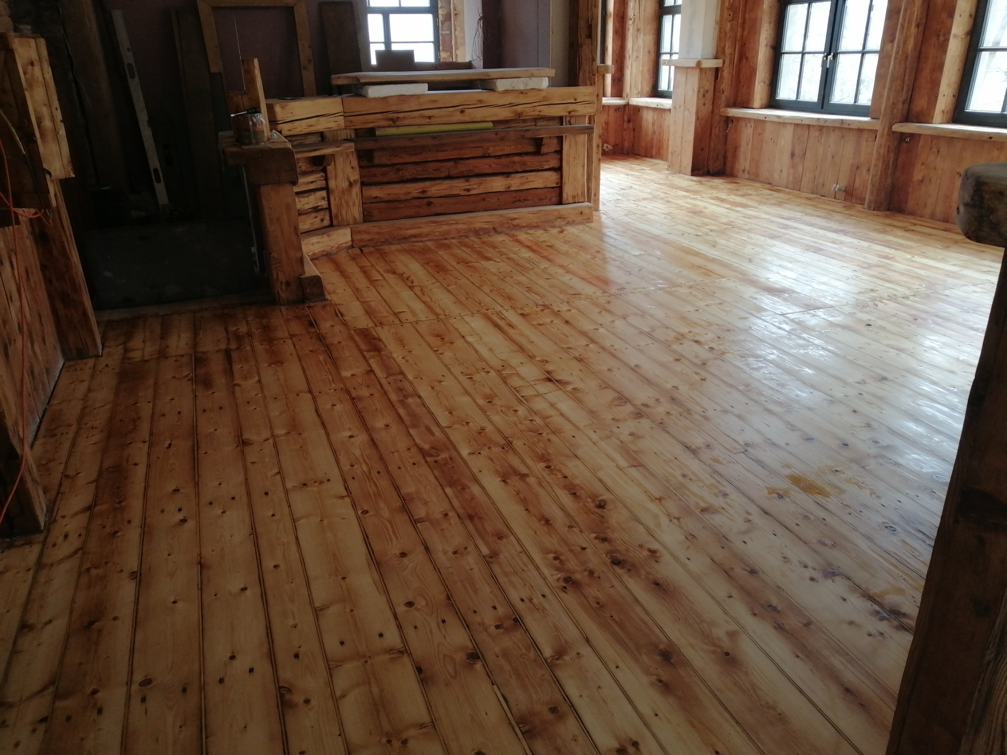 Świeżo polakierowana, jasna podłoga z desek w rustykalnym wnętrzu, widoczne drewniane ściany i okna z czarnymi ramami.