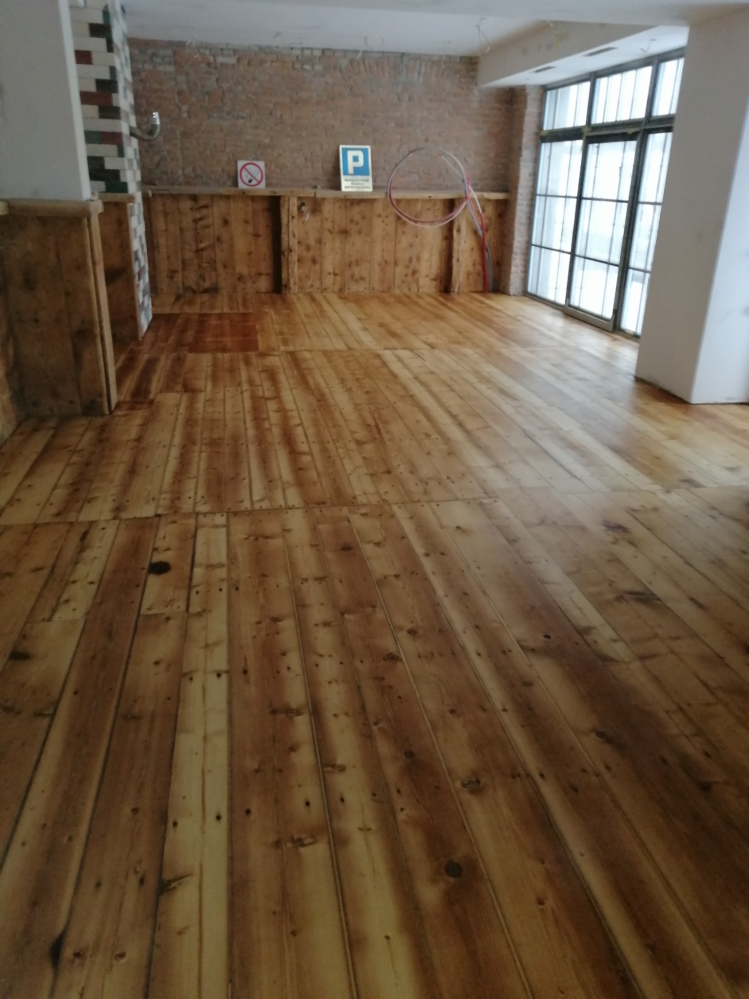 Wykończona podłoga z desek w pomieszczeniu z surową cegłą na ścianie, widoczne ślady po obróbce i niedoskonałości naturalnego drewna, duże okno z czarnymi szprosami w tle.