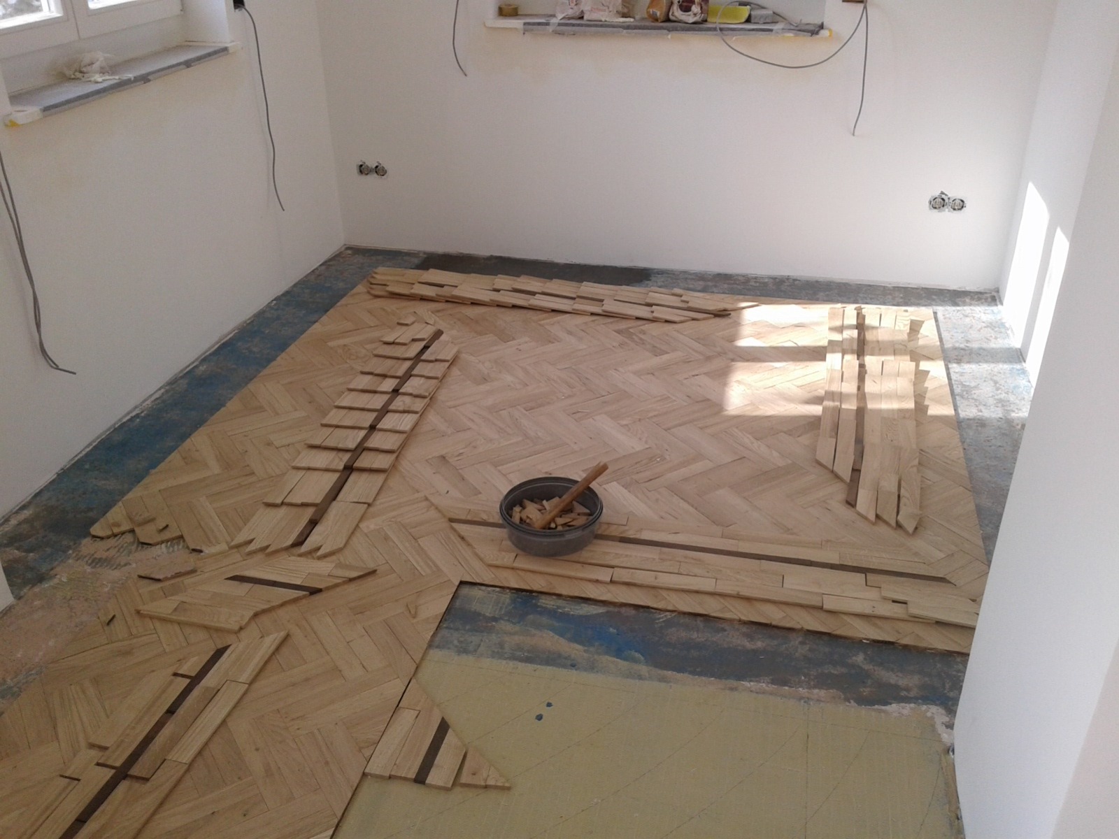 Układanie parkietu w jodełkę, widoczny fragment podłogi z przygotowanymi klepkami, wiaderko z narzędziami i klejem, surowe ściany w tle.