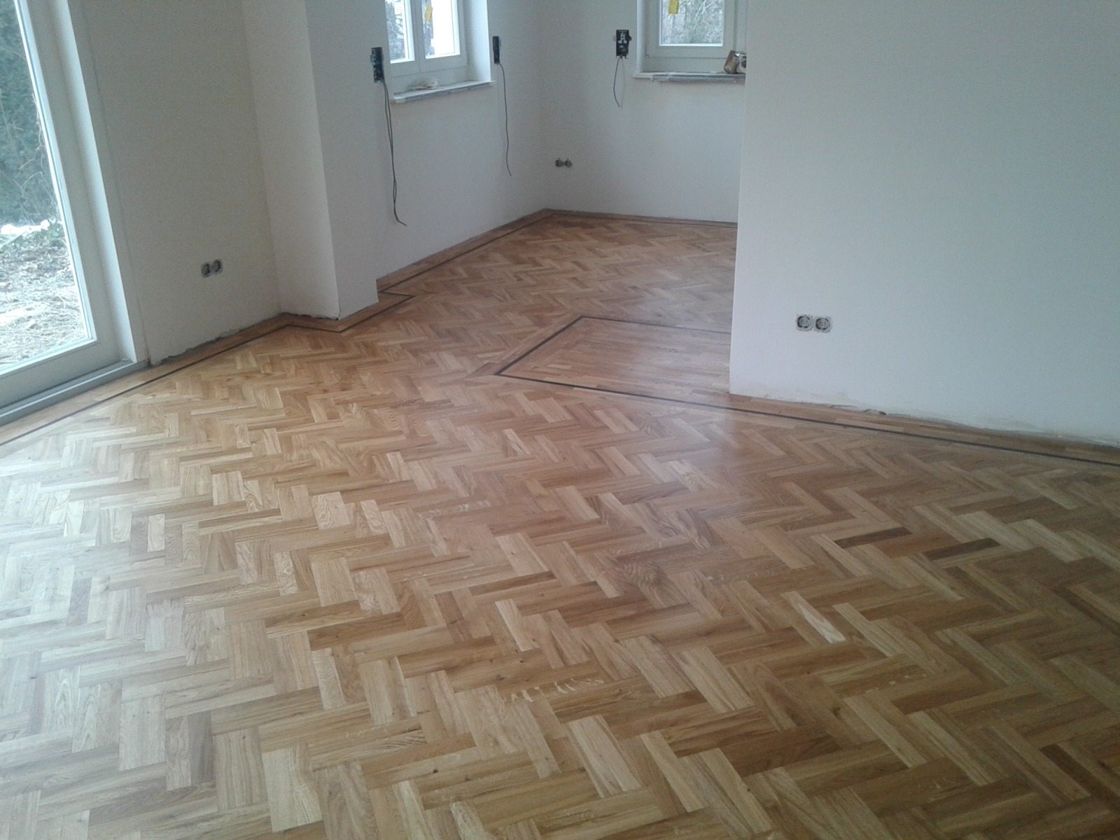 Ułożony parkiet w jodełkę klasyczną, z ciemniejszym obramowaniem przy ścianach i w formie kwadratu na środku, widoczne gniazdka elektryczne na ścianach, okna w tle.