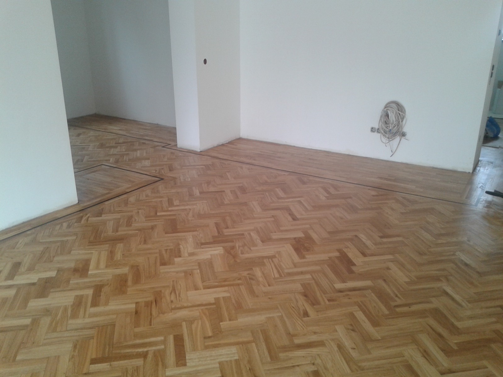 Ułożony parkiet w jodełkę w jasnym odcieniu drewna, z ciemnymi listwami przypodłogowymi i widocznymi kablami wystającymi ze ściany.