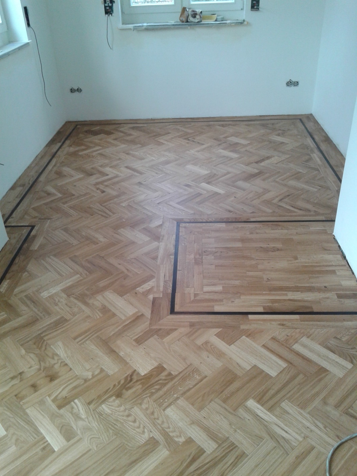 Nowa, jasna podłoga parkietowa ułożona we wzór jodełki francuskiej z ciemną bordiurą w pomieszczeniu z białymi ścianami i oknem.