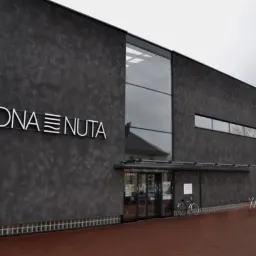 Elewacja budynku z ciemnoszarą, chropowatą fakturą, z dużym przeszkleniem i białym napisem 'WODNA NUTA' nad wejściem, rower oparty o ścianę.