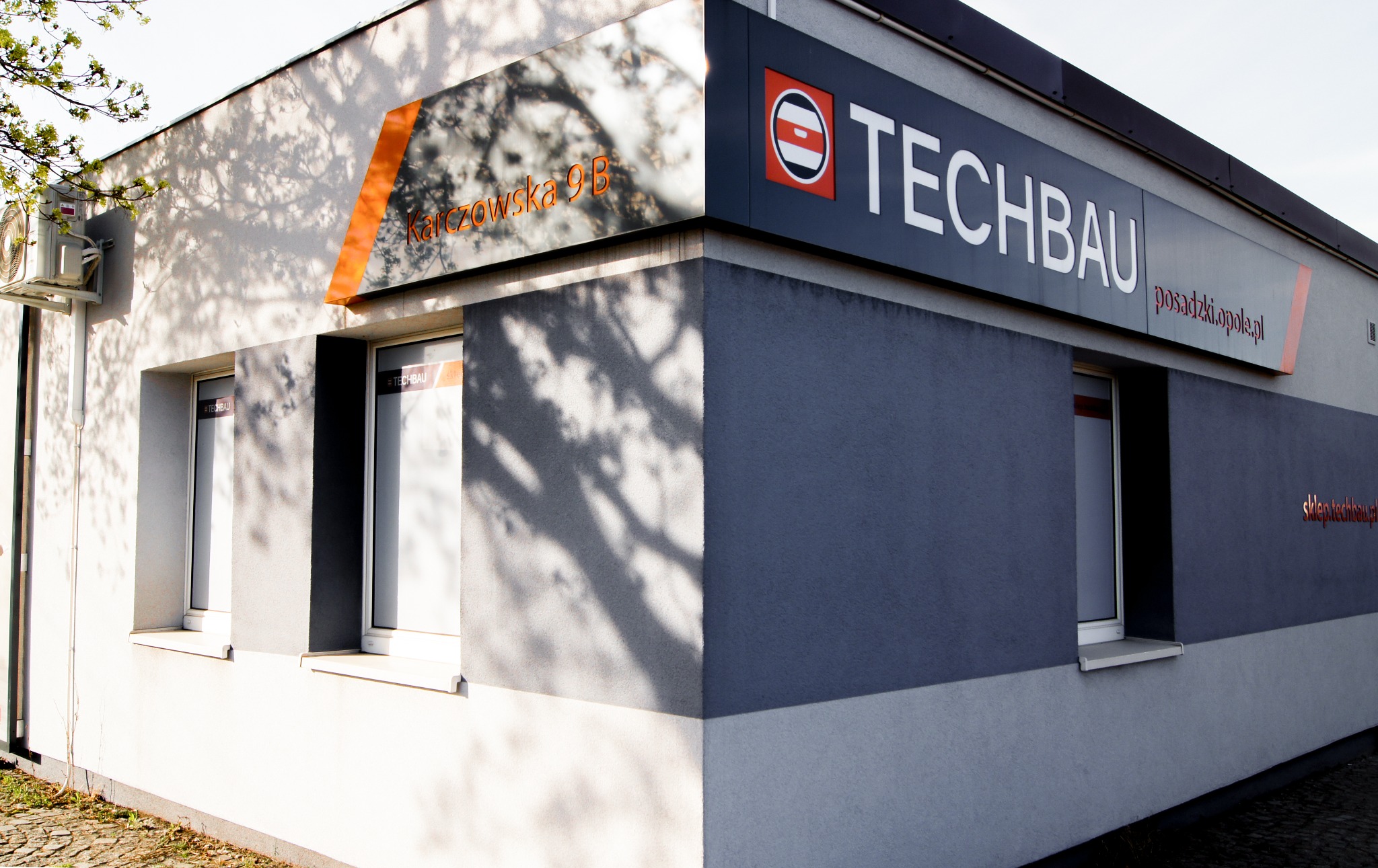 Zewnętrzny widok narożnika budynku firmy TECHBAU z dwoma oknami, klimatyzatorem i szyldem z logo oraz adresem Karczowska 9B, z cieniami drzew na elewacji.