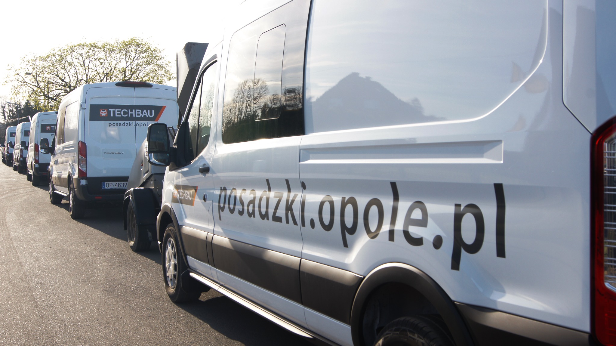 Flota białych samochodów dostawczych firmy Techbau z Opola, specjalizującej się w posadzkach, zaparkowana w rzędzie na tle drzew.