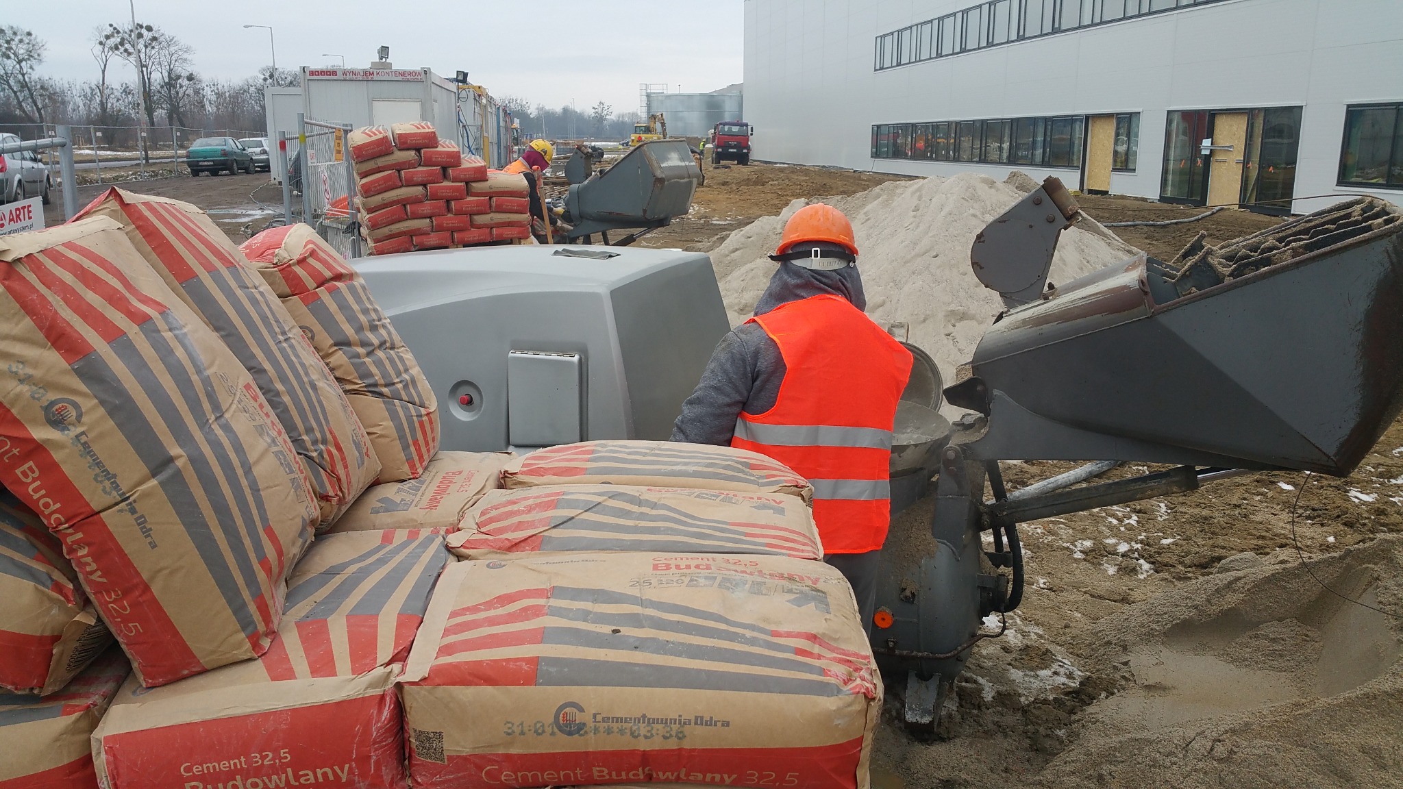 Pracownik w pomarańczowej kamizelce obsługuje maszynę do mieszania zaprawy obok stosu worków z cementem budowlanym 32,5 na placu budowy, w tle widoczny nowy budynek.