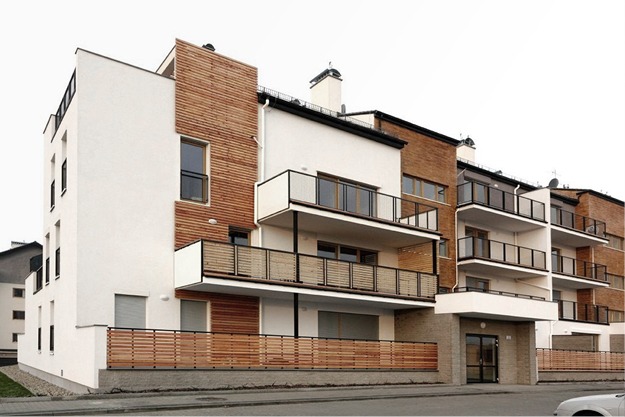 Nowoczesny budynek apartamentowy z balkonami i elewacją łączącą biały tynk z drewnianymi panelami, widziany z poziomu ulicy.