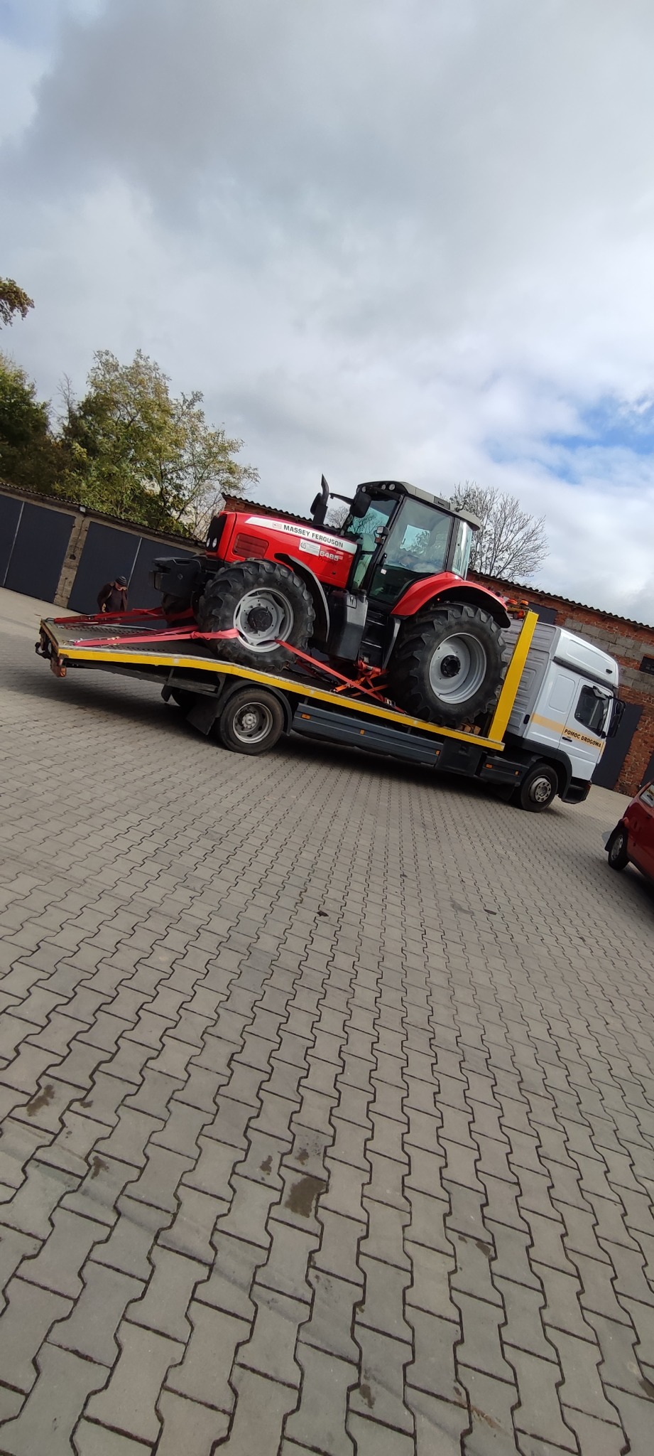 Czerwony traktor Massey Ferguson zabezpieczony pasami na żółtej platformie lawety z białą kabiną, na tle szarej kostki brukowej i pochmurnego nieba.