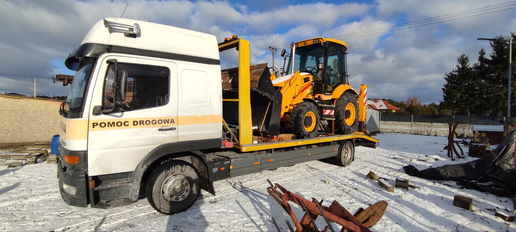 Żółta koparko-ładowarka JCB przewożona na lawecie pomocy drogowej z napisem 'POMOC DROGOWA', zimowy krajobraz w tle.