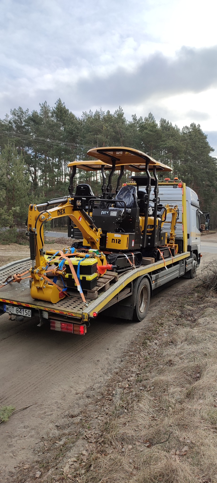 Dwie mini koparki NEX N12 zabezpieczone pasami na platformie lawety, widok z tyłu, tablica rejestracyjna WOT 84150.