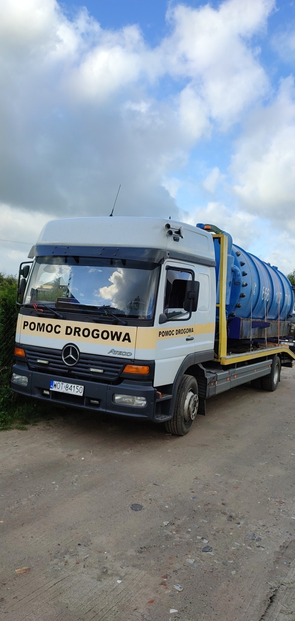 Laweta Mercedes-Benz Atego z niebieską cysterną na platformie, oznaczona jako Pomoc Drogowa, zaparkowana na żwirowej drodze pod pochmurnym niebem.