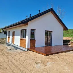 Nowoczesny parterowy dom z białym tynkiem, czarnym dachem i drewnianymi elementami wykończeniowymi, z przylegającym tarasem z ciemnego drewna, w słoneczny dzień.