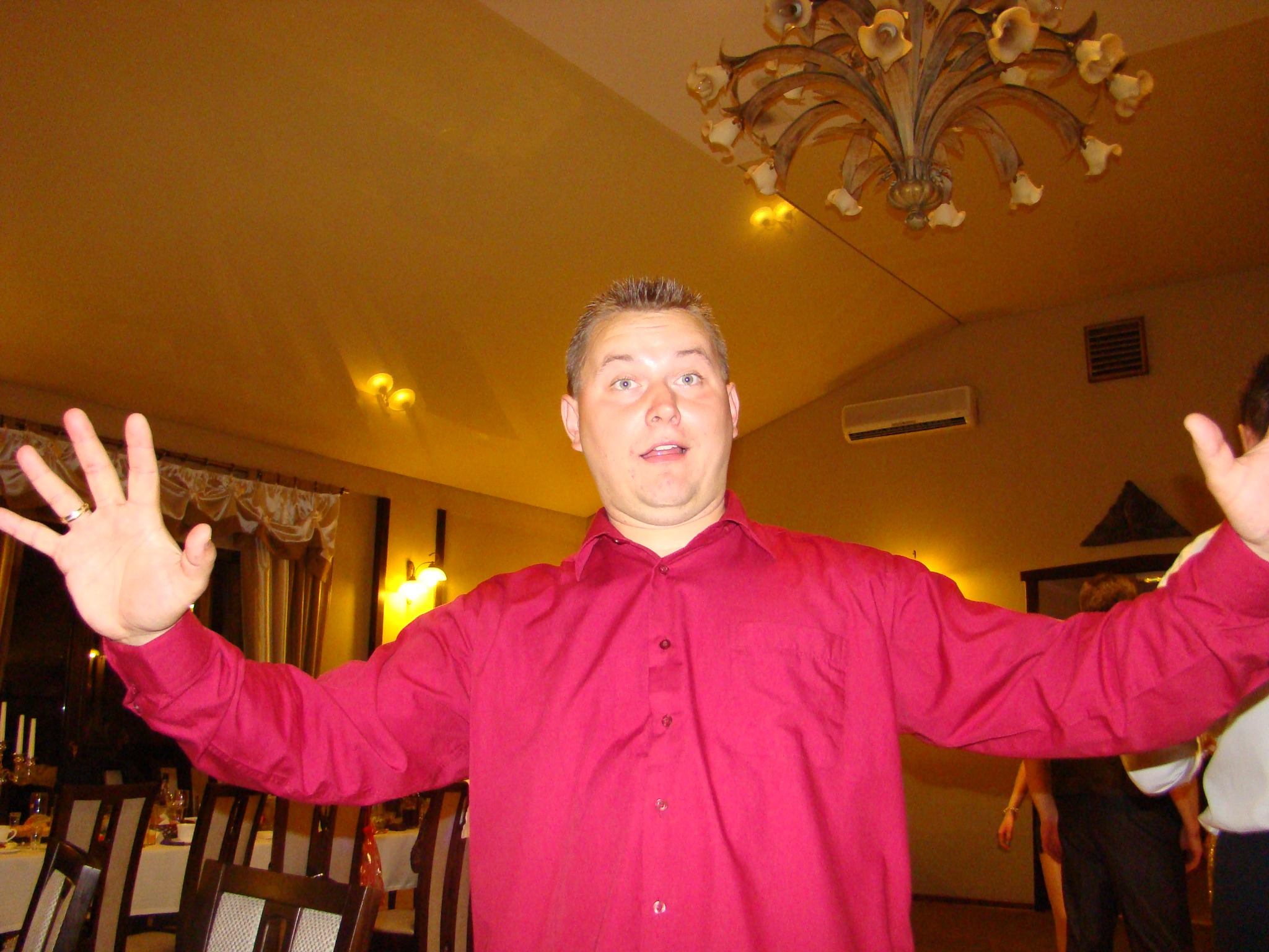 Mężczyzna w czerwonej koszuli z rozłożonymi rękami w sali z dekoracyjnym żyrandolem i stołami.