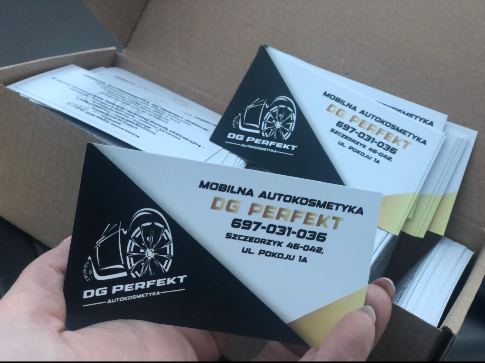 Wizytówki firmy DG Perfekt Autokosmetyka, z logo przedstawiającym zarys samochodu i koła, ułożone w kartonowym pudełku, trzymane w dłoni.