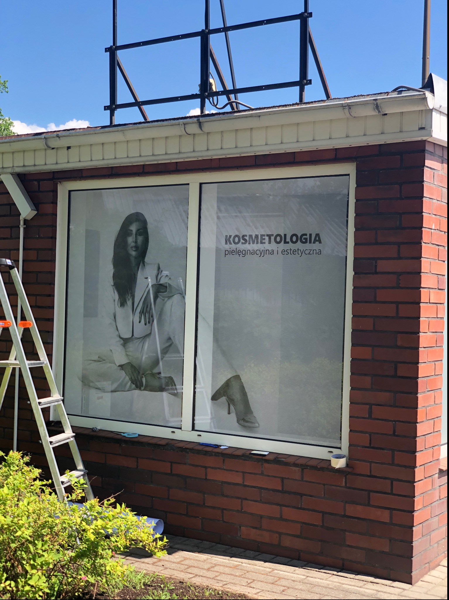 Oklejone folią okno z reklamą salonu kosmetycznego, drabina oparta o ścianę budynku z czerwonej cegły, częściowo widoczny zwój folii i nożyczki.