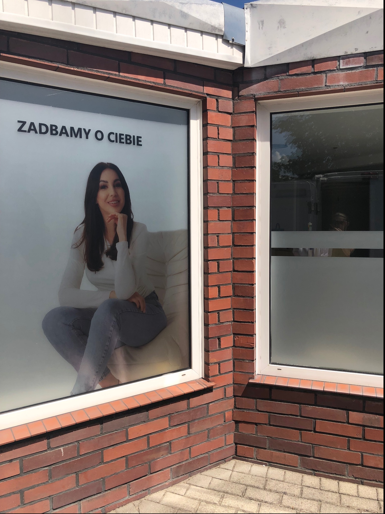 Oklejona witryna salonu z wizerunkiem uśmiechniętej kobiety i napisem 'Zadbamy o Ciebie', widoczna ceglana elewacja budynku.
