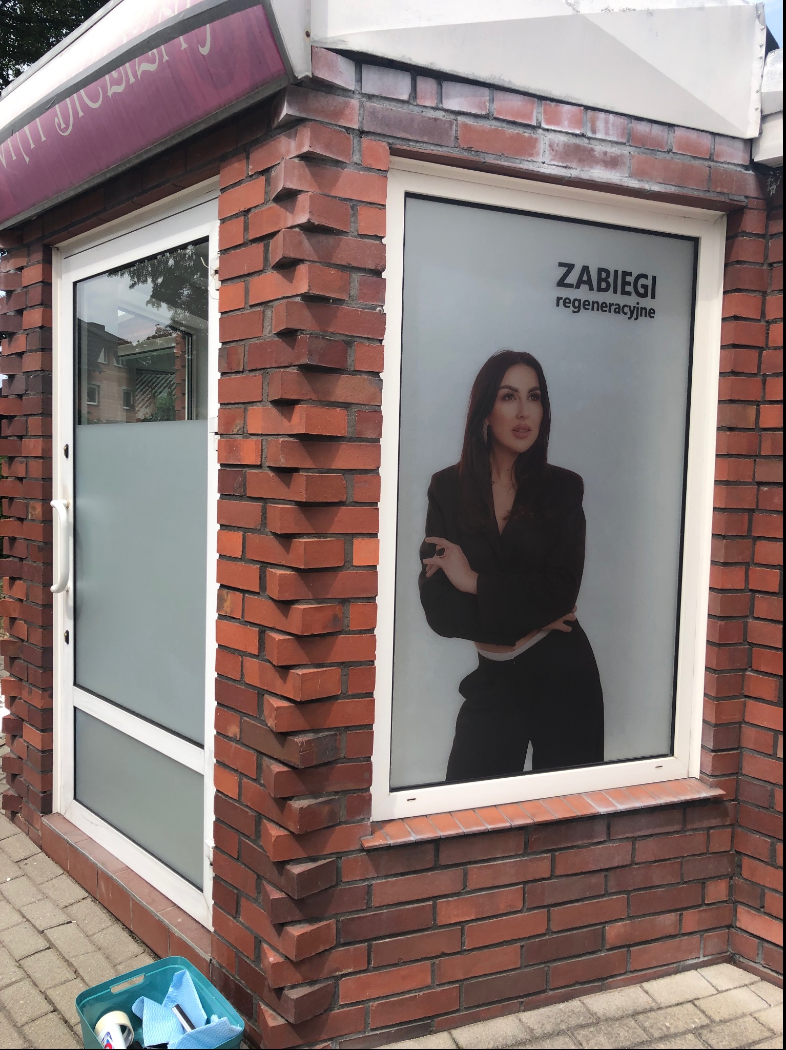 Oklejone okno salonu kosmetycznego z wizerunkiem kobiety i napisem 'Zabiegi regeneracyjne', widoczne narzędzia w pojemniku obok.