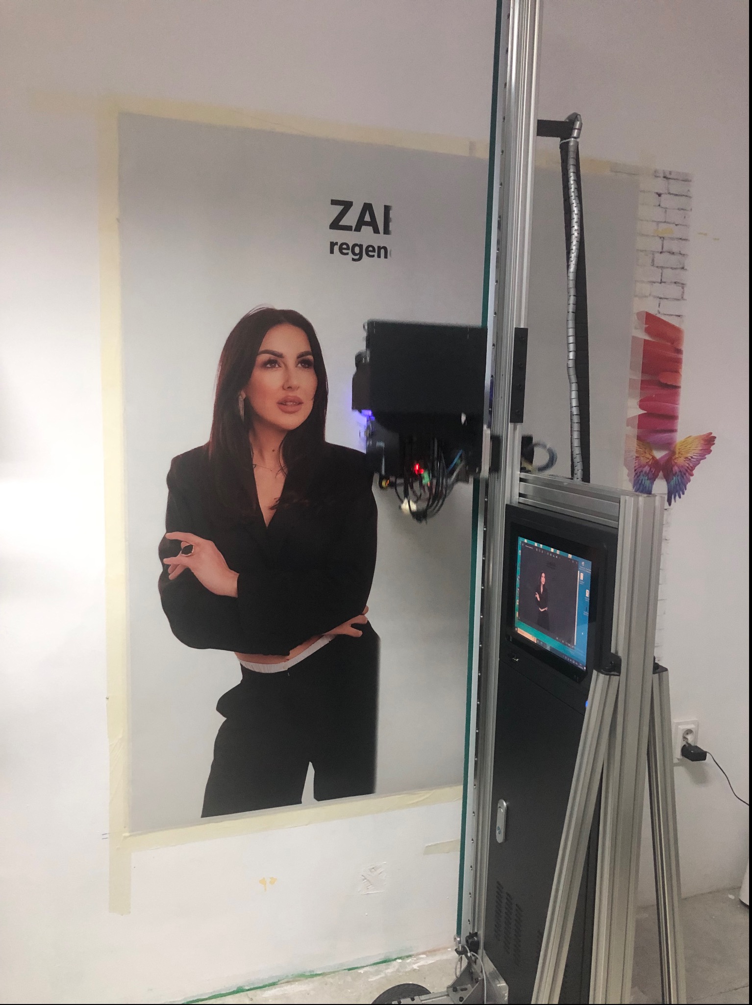 Drukarka pionowa drukująca portret kobiety na ścianie, widoczne kable, monitor drukarki i taśma malarska zabezpieczająca krawędzie druku.
