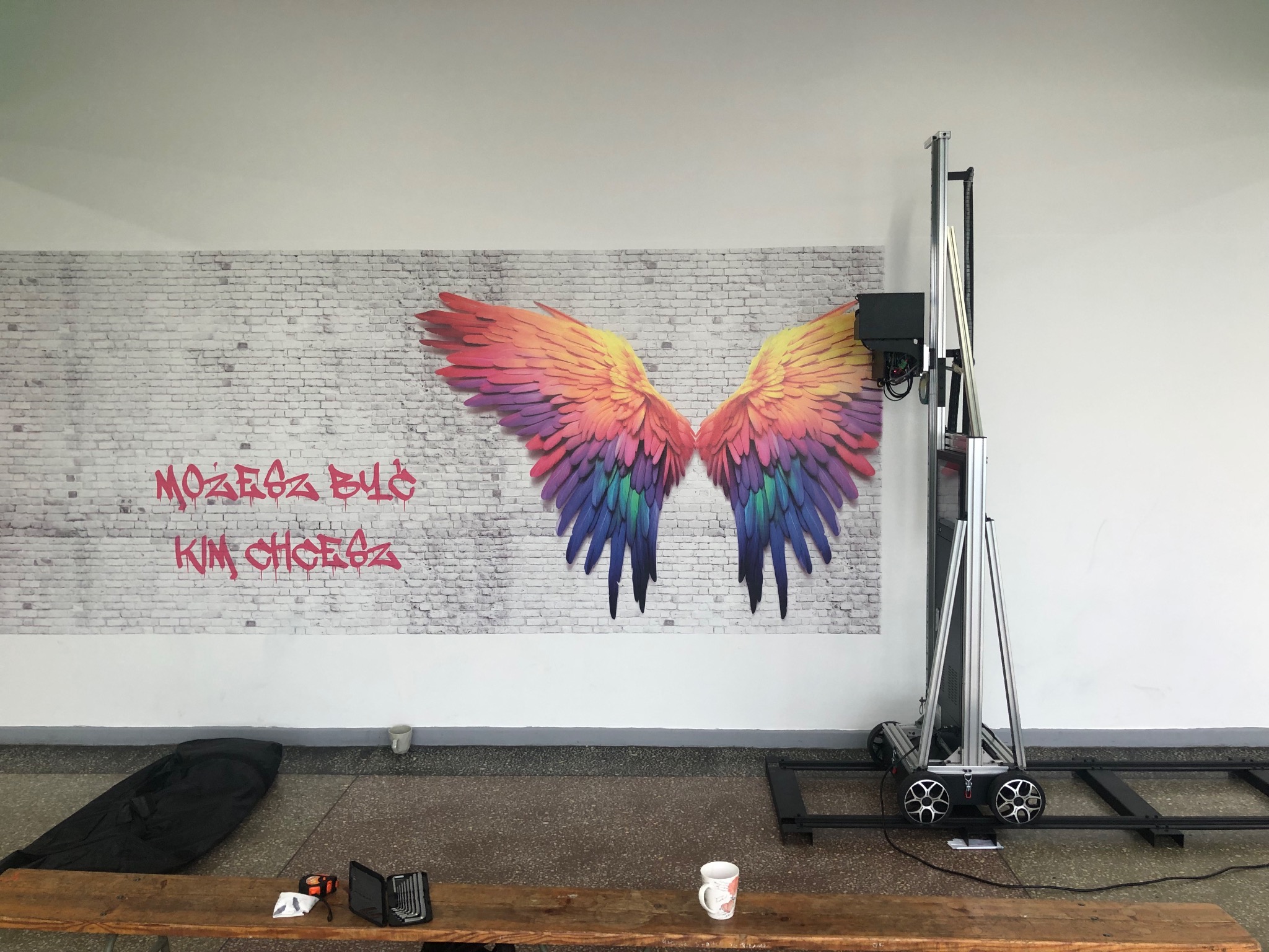 Ściana z nadrukowanymi kolorowymi skrzydłami i napisem graffiti, obok stoi pionowa drukarka do ścian.