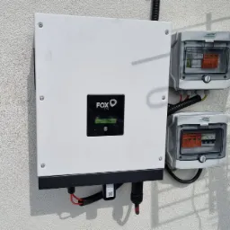 Biała obudowa inwertera Fox ESS zamontowana na chropowatej ścianie zewnętrznej, z widocznymi dwoma szarymi skrzynkami zabezpieczeń z przezroczystymi pokrywami i przewodami w peszlach.