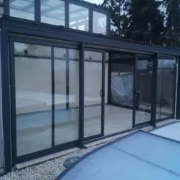 Zadaszenie basenowe z przesuwnymi, aluminiowymi ramami szklanymi, widok z zewnątrz, częściowo zasłonięty basen. Widoczne drzewa w tle.