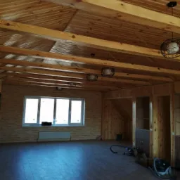 Surowe wnętrze z drewnianym sufitem, ścianami z boazerii i wykończoną podłogą, widoczne lampy wiszące i okno z grzejnikiem, sprzęt do sprzątania w rogu.