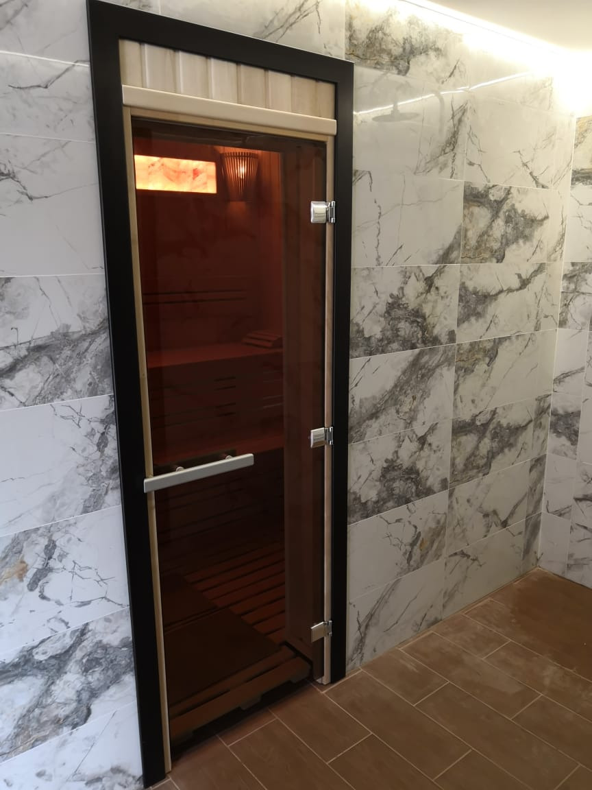 Szklane drzwi do sauny z drewnianym wykończeniem, widoczne marmurowe płytki na ścianie i brązowa podłoga z płytek.