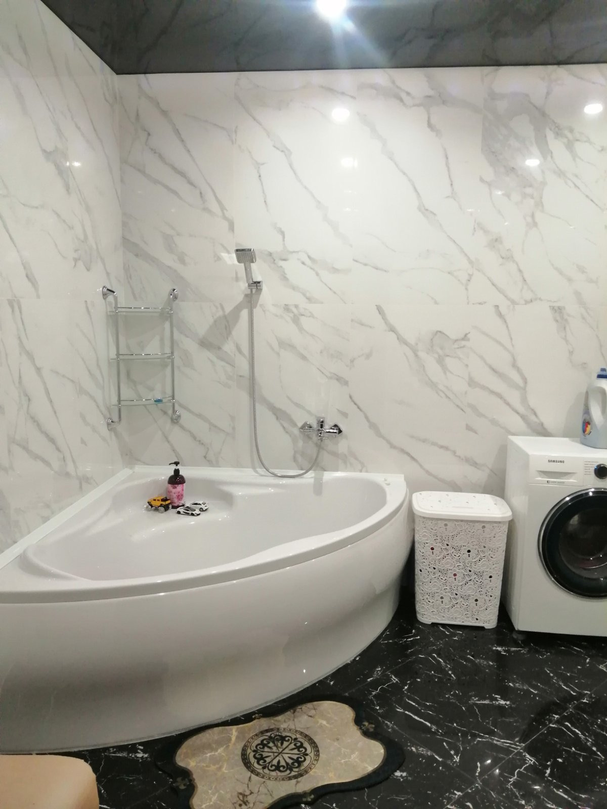 Nowoczesna łazienka z narożną wanną, wykończona białymi płytkami imitującymi marmur i czarną marmurową podłogą. Na wannie stoją zabawki, a obok wanny znajduje się pralka i ażurowy kosz na pranie.