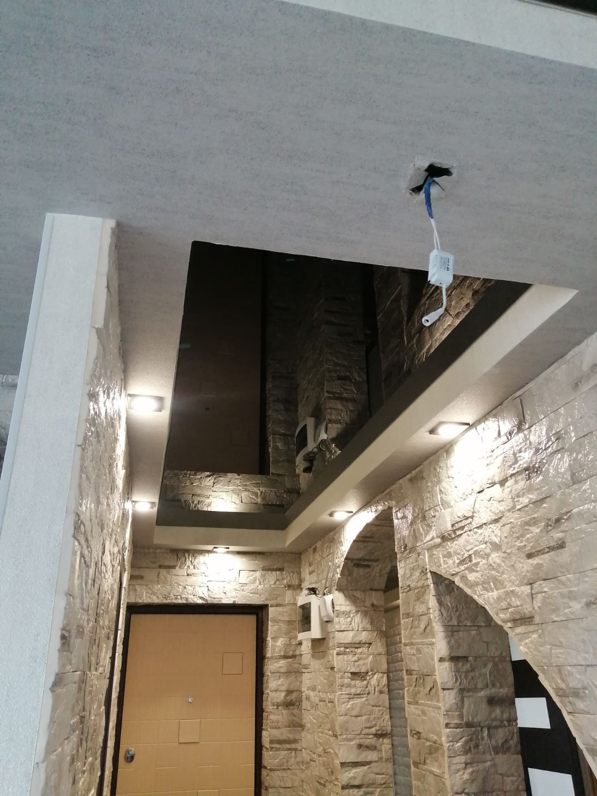 Korytarz po remoncie z dekoracyjną ścianą z kamienia, oświetleniem punktowym w suficie podwieszanym i widocznymi przewodami elektrycznymi w miejscu montażu lampy.