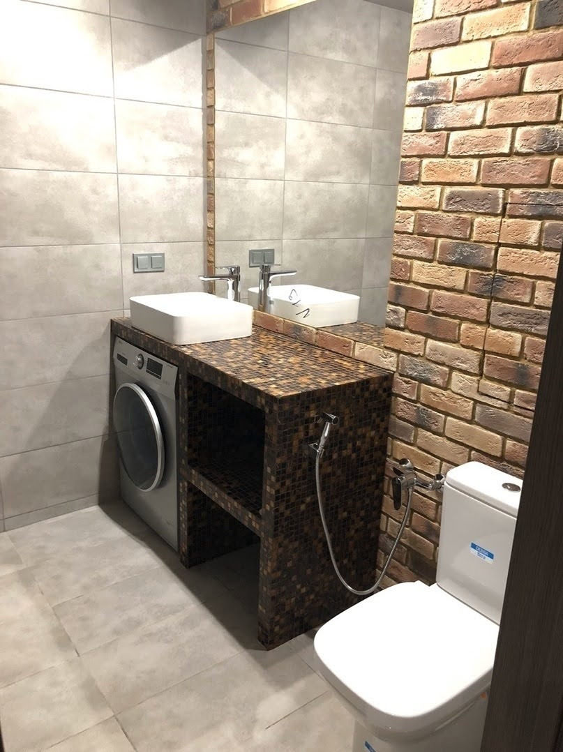 Nowoczesna łazienka z umywalkami nablatowymi, pralką ukrytą pod blatem z mozaiki, ścianą z cegły i szarą płytką oraz toaletą z bidetką.