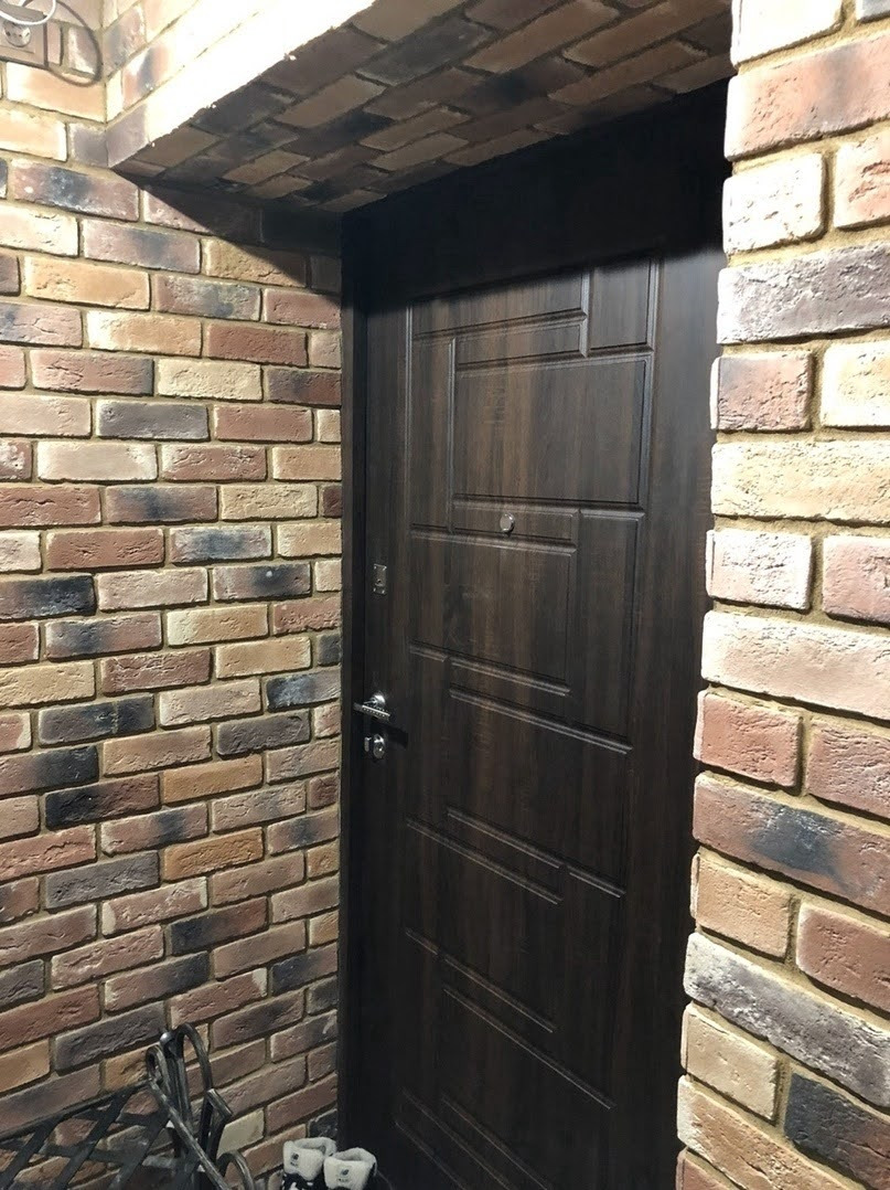 Ciemne, drewnopodobne drzwi wejściowe w otworze obłożonym dekoracyjną cegłą w odcieniach brązu i beżu.