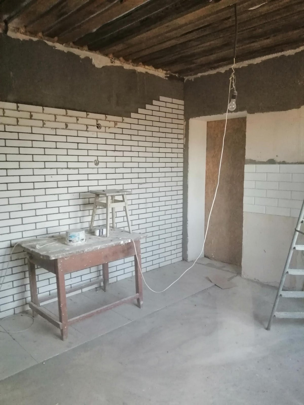 Wnętrze w trakcie prac: częściowo ułożona biała cegiełka na ścianie, surowy sufit z drewnianych belek, prowizoryczne oświetlenie żarówką na kablu, drewniany stół z puszką i drabina w tle.