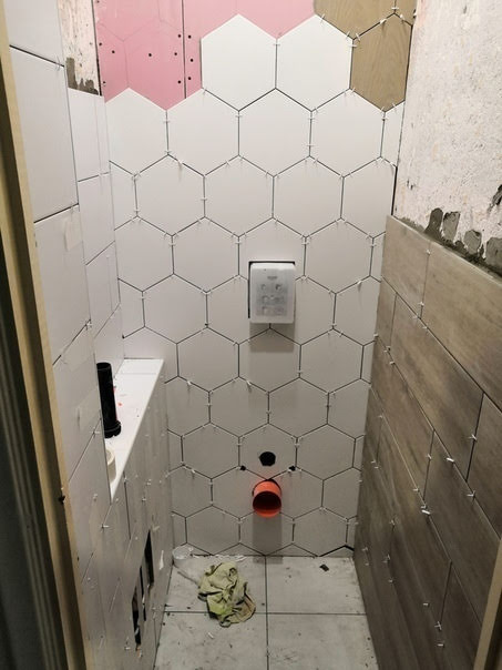 Układanie białych, sześciokątnych płytek w łazience, widoczne krzyżyki dystansowe, fragment ściany z płytą kartonowo-gipsową i instalacją hydrauliczną.