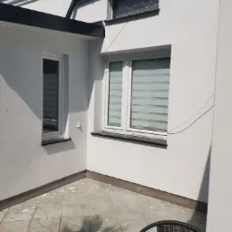 Świeżo pomalowana, biała elewacja narożna budynku z dwoma oknami z roletami dzień-noc, widoczna płytka cokołowa i fragment tarasu z szarych płytek.