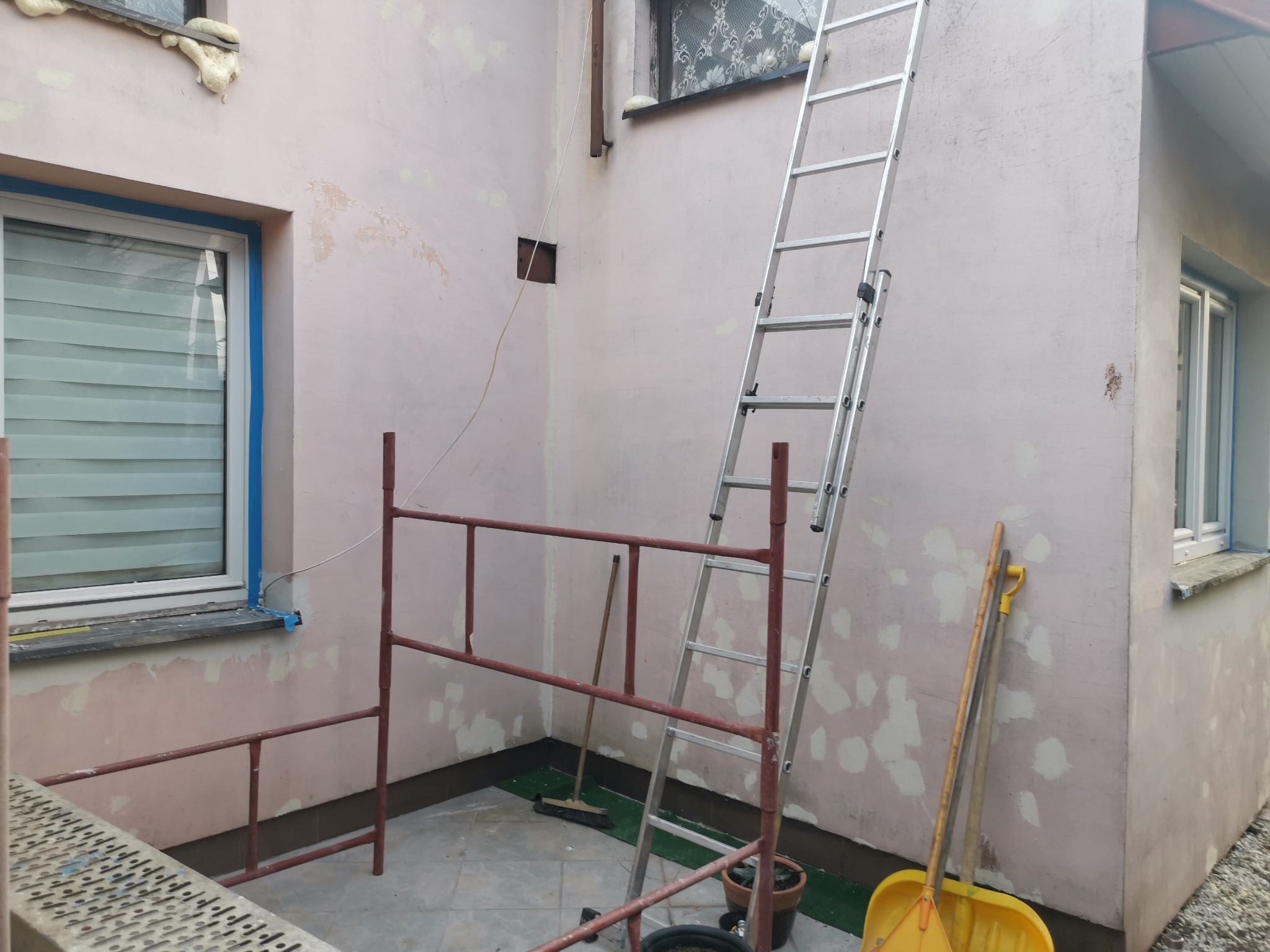Przygotowania do prac na elewacji: ściana z nałożonymi łatami, okna zabezpieczone taśmą, rozstawiony ruszt i drabina oparta o ścianę, widoczne narzędzia.