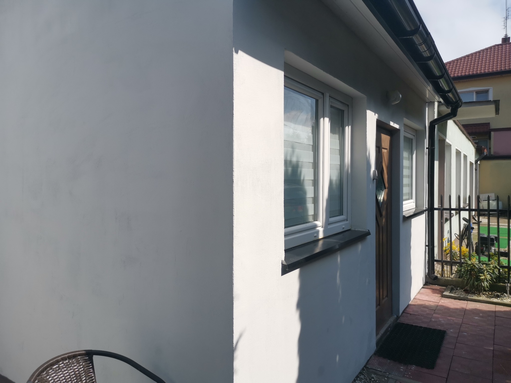 Świeżo pomalowana, biała elewacja domu jednorodzinnego z widocznym oknem z żaluzjami, ciemną rynną i fragmentem chodnika z kostki.