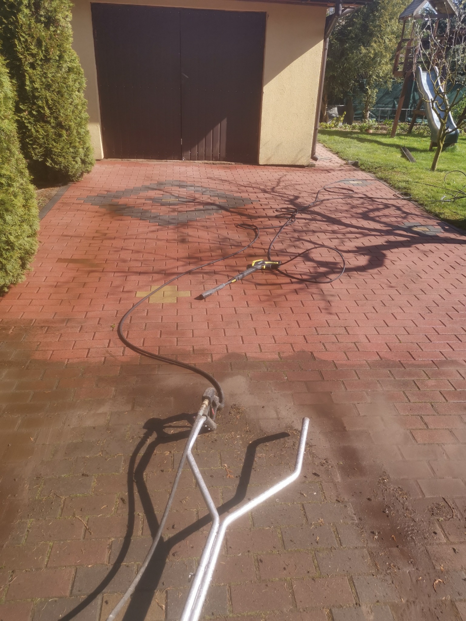 Czerwona kostka brukowa w trakcie czyszczenia myjką ciśnieniową; widoczne różnice w kolorze między czystą i zabrudzoną powierzchnią, w tle garaż i huśtawka.