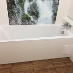 Biała wanna obudowana płytkami z motywem wodospadu na ścianie, drewnopodobna podłoga i chromowana bateria.