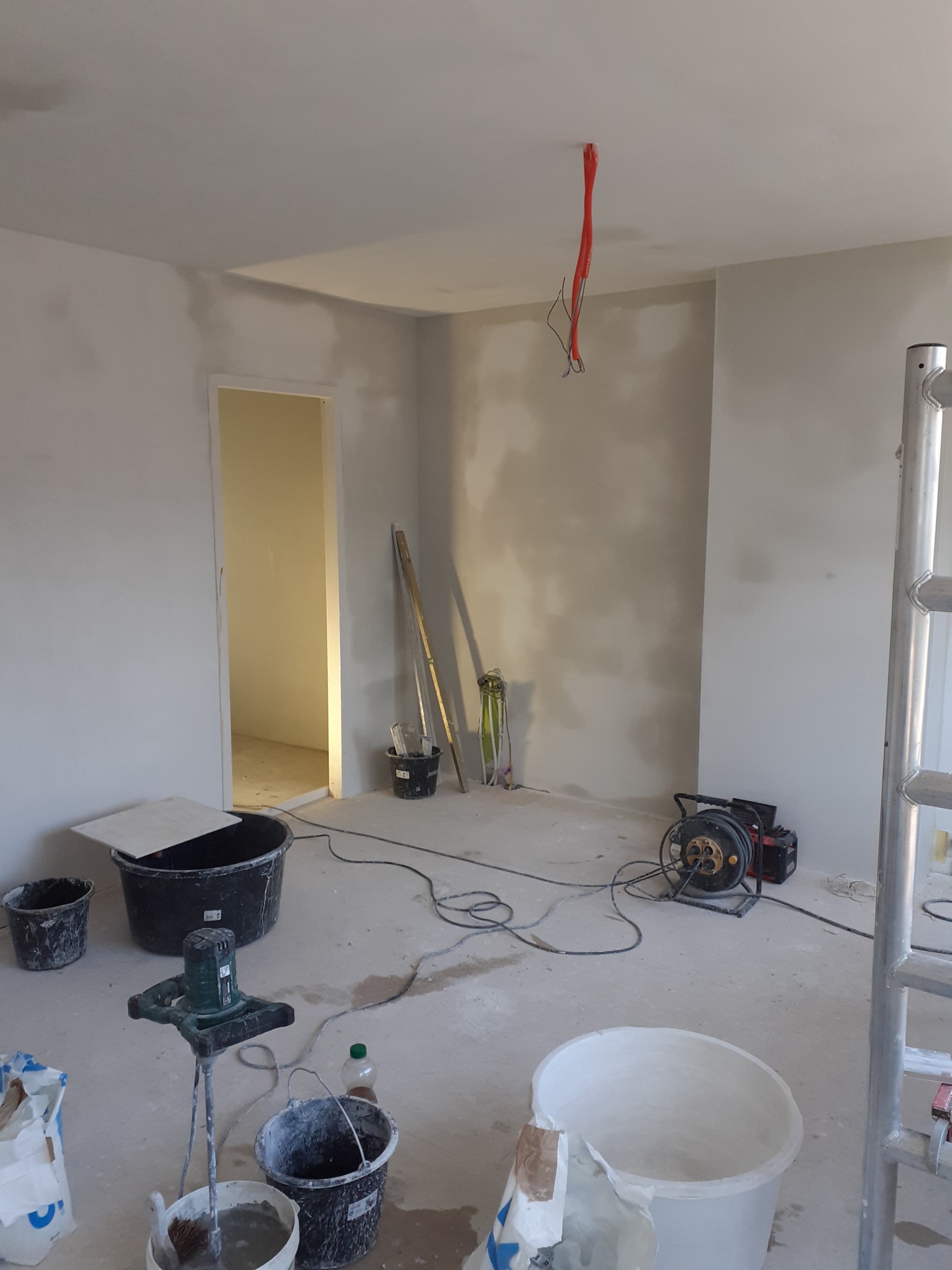 Wnętrze pokoju w trakcie prac wykończeniowych: ściany po nałożeniu gładzi, widoczne narzędzia, wiadro z zaprawą, mieszadło elektryczne, przedłużacz zwinięty na bębnie i wystające przewody...