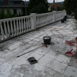 Taras w trakcie demontażu płytek, widoczne narzędzia: łopata, miotła, wiadro z gruzem, czerwony kabel, w tle biała balustrada i budynki.