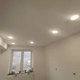 Wnętrze świeżo pomalowanego pokoju z trzema okrągłymi lampami halogenowymi w suficie, widok na okno z parapetem zastawionym różnymi przedmiotami, grzejnik pod oknem, ślady po gniazdkach na ścianie...